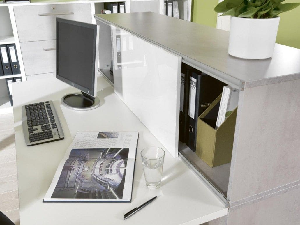 Arbeitsplatz, in hellgrau und weiß, ein PC steht darauf, daneben liegt ein offenes Prospekt