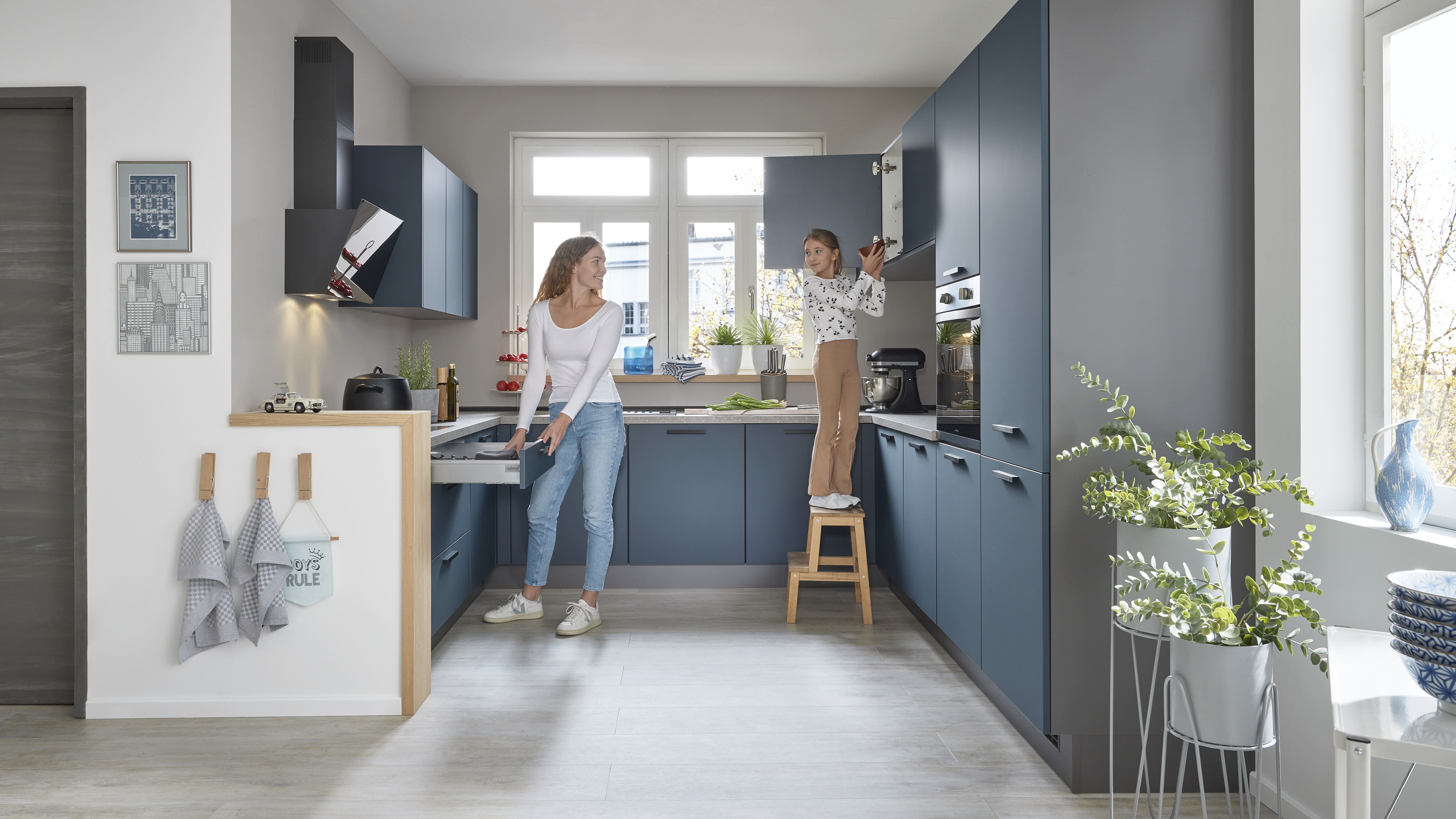 Hellgraue Küche im matten Stil, eine Frau räumt etwas in eine Schublade und ein Mädchen steht auf einem Hocker, um etwas aus einem Schrank zu holen.