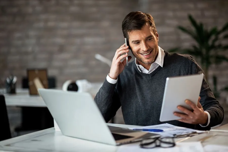 Mann mit Tablet in der Hand, vor ihm ein Laptop. Er ist am telefonieren
