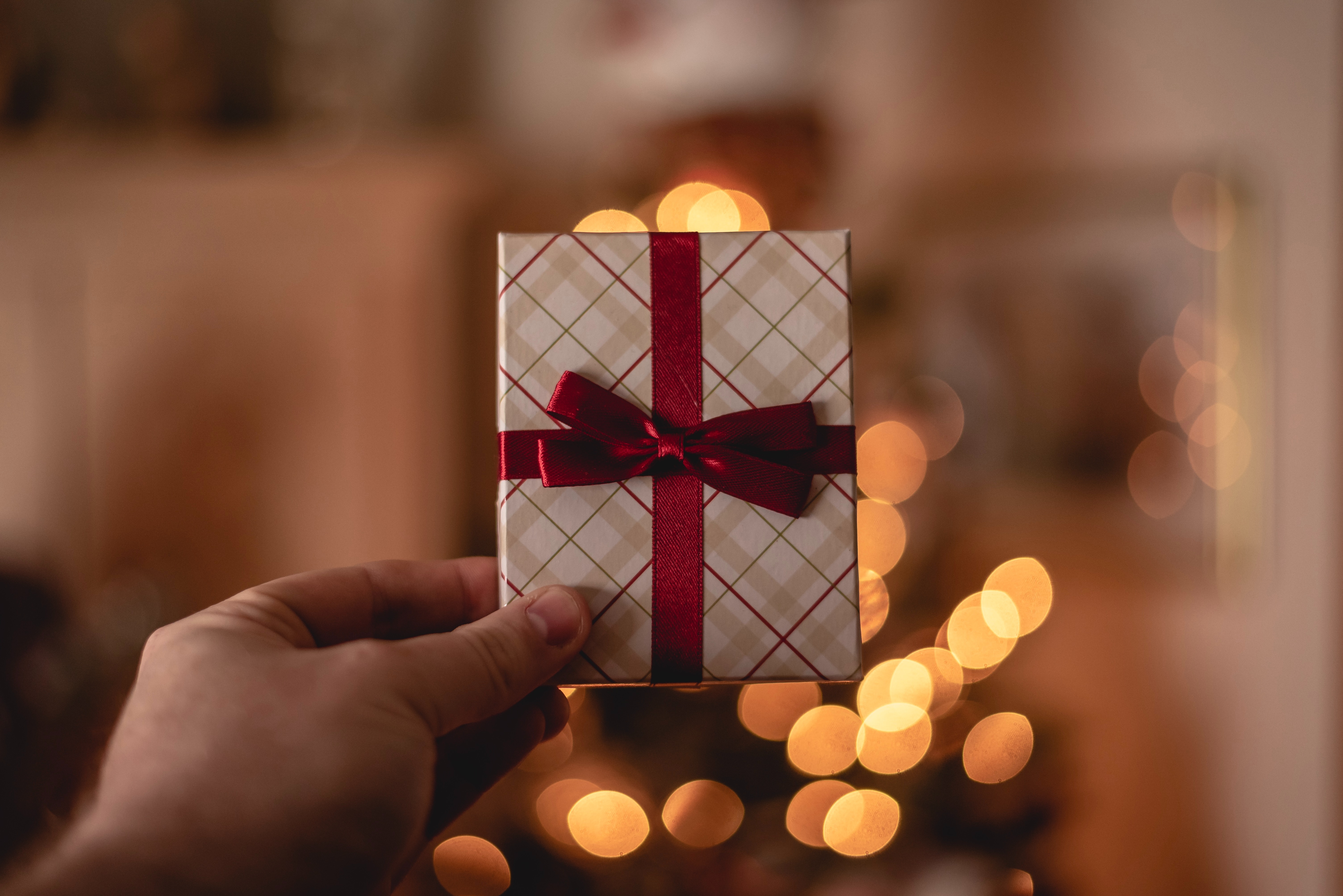 kleines Geschenk, das hochgehalten wird; im Hintergrund sieht man verschwommen die Lichter eines Christbaumes