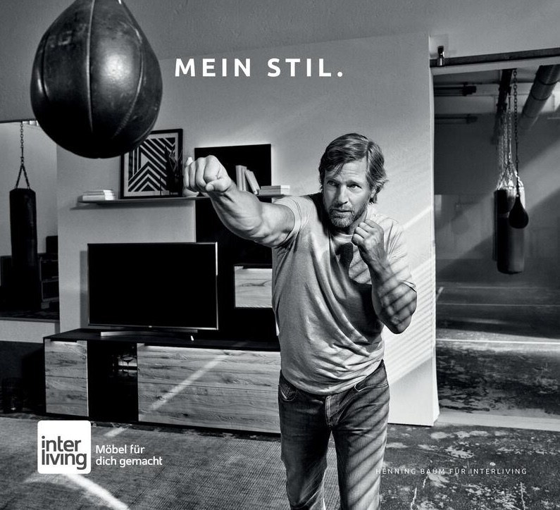 Schwarz-Weiß Bild eines Mannes, der gegen einen Boxsack boxt, hinter ihm eine moderne Wohnwand