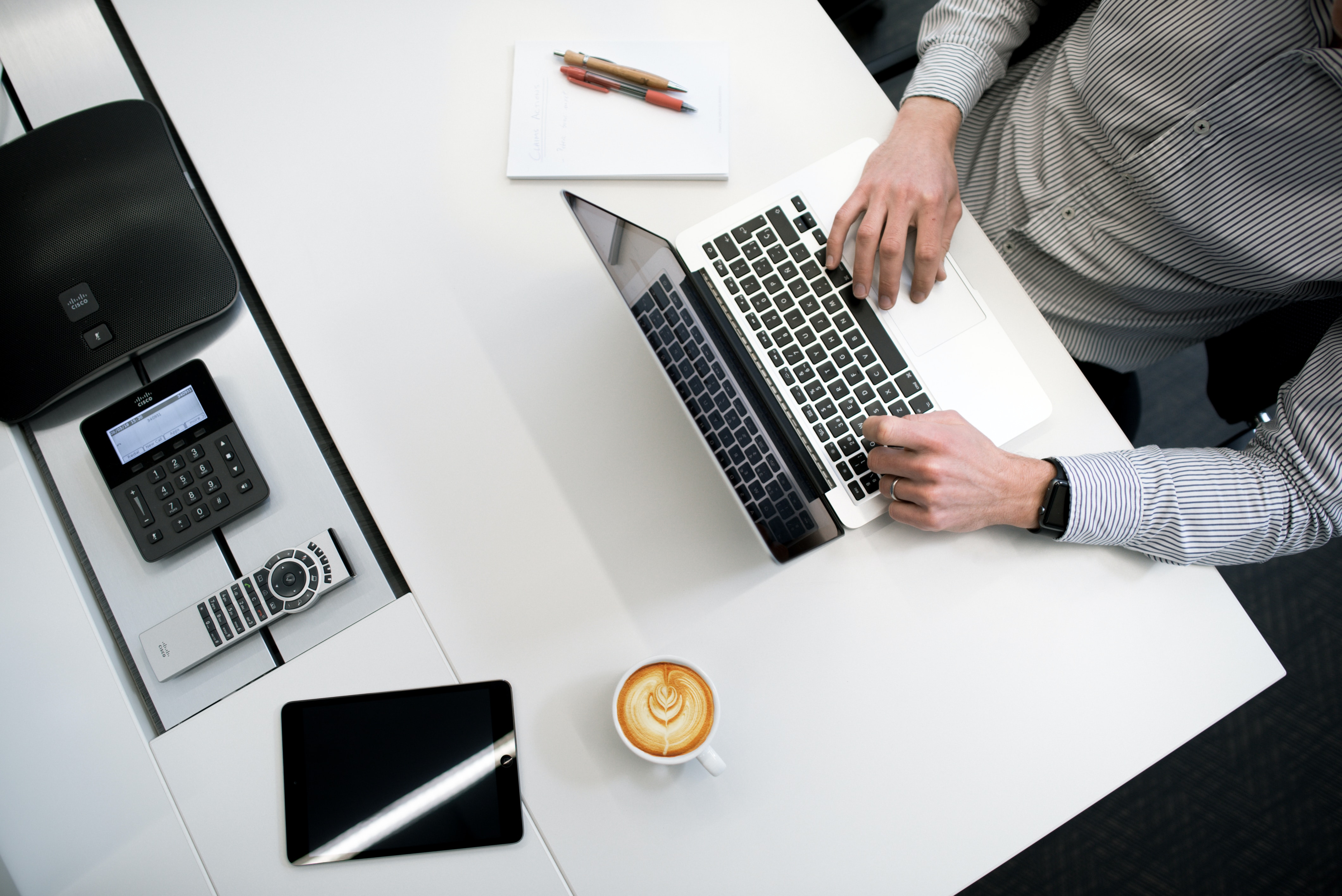 Ansicht von oben auf einen Schreibtisch. Dort steht ein Kaffee und ein Laptop, an dem ein Mann in einem grauen Hemd sitzt