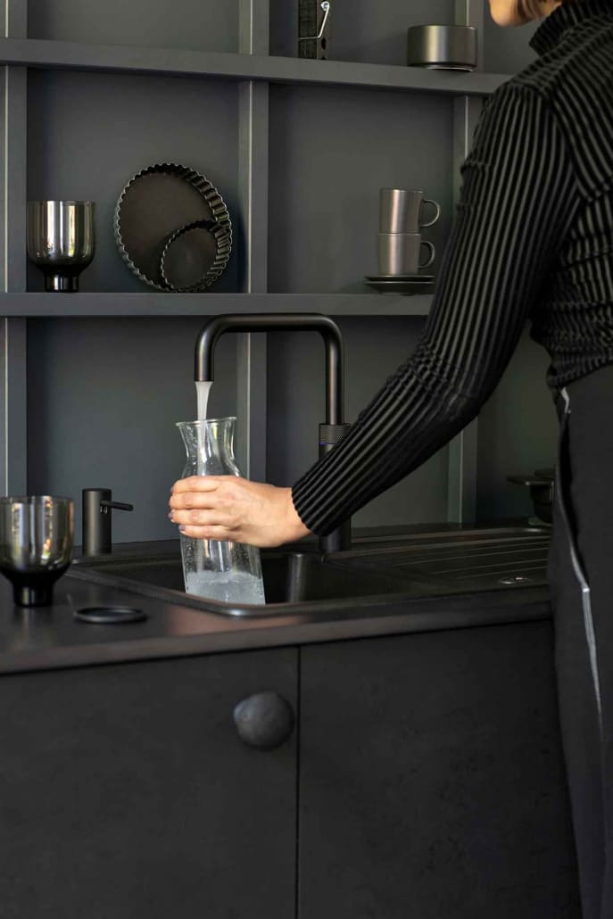 Schwarzer Quooker an einem schwarzen Waschbecken, eine Person füllt dort gerade ein Wasserglas auf