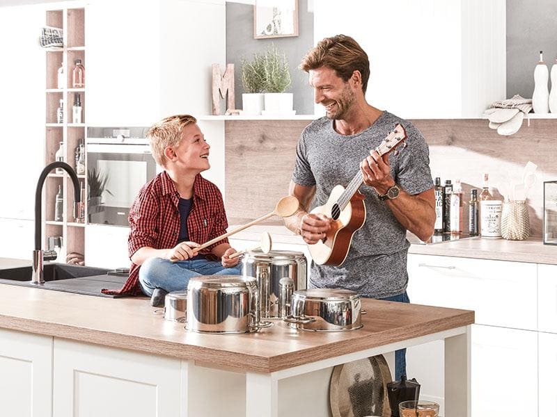Weiße Küche mit hellen Holzablagen, auf einer Ablage sitzt ein Junge, der lacht, daneben steht sein Vater, der Gitarre spielt und ebenso lacht