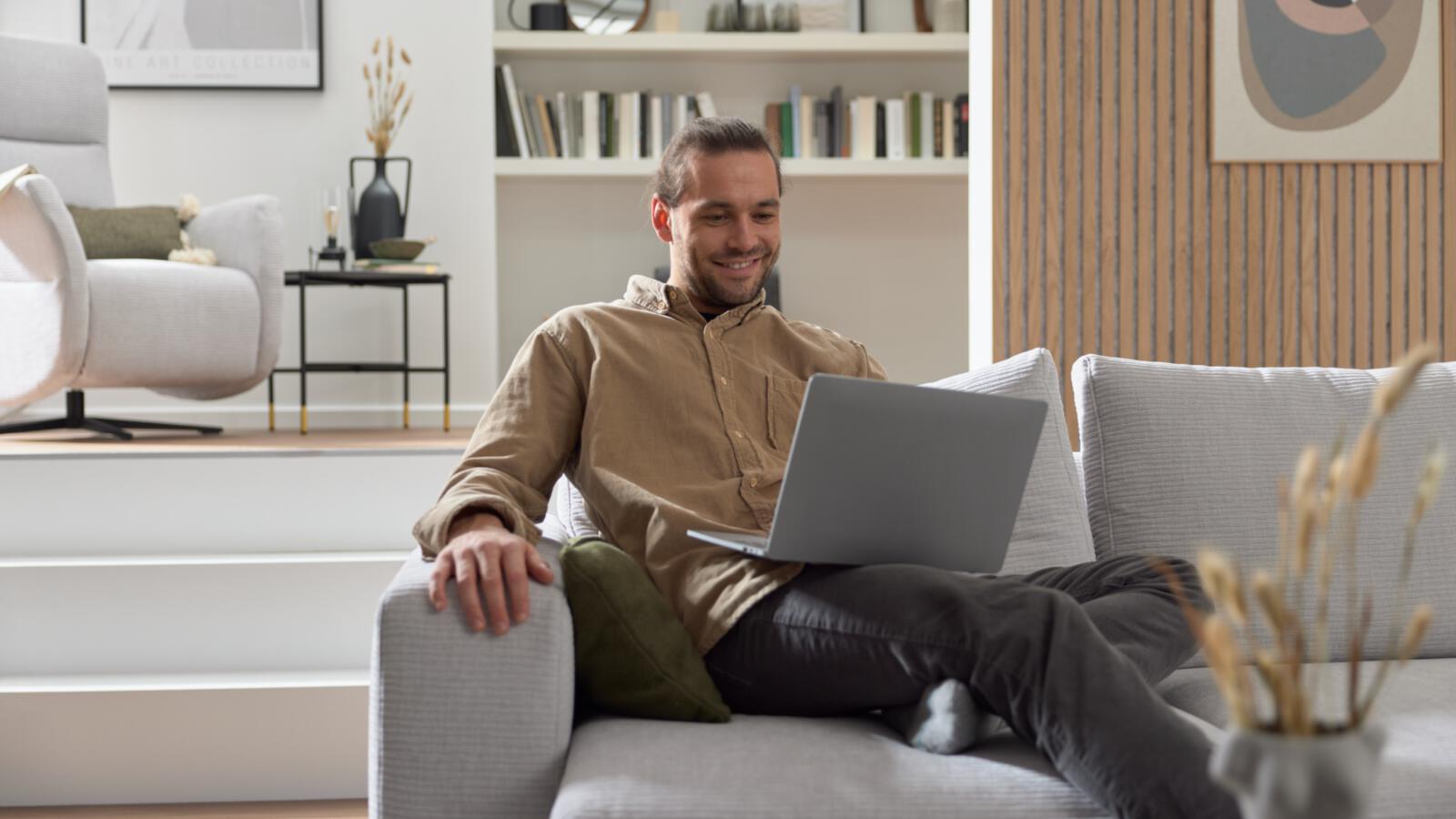 Mann sitzt auf einem grauen Sofa und hat einen Laptop auf dem Schoß, er lächelt