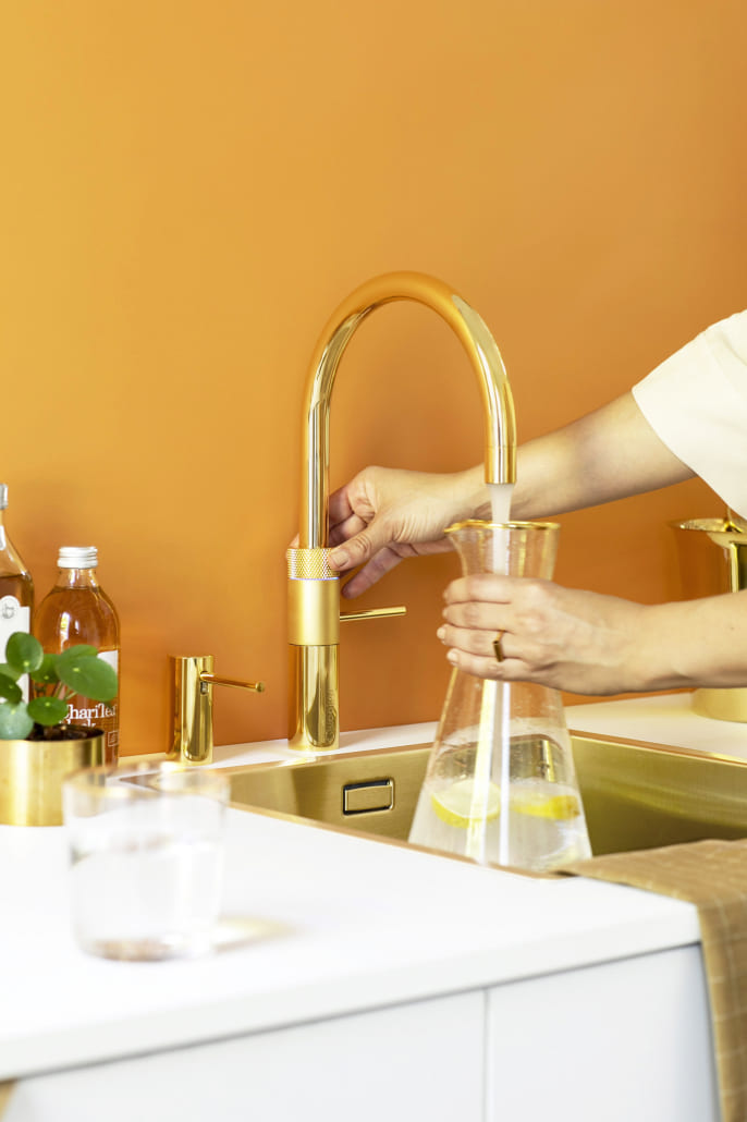 Goldener Quooker vor einer gelben Wand, eine Frau füllt dort gerade ihre Karaffe auf