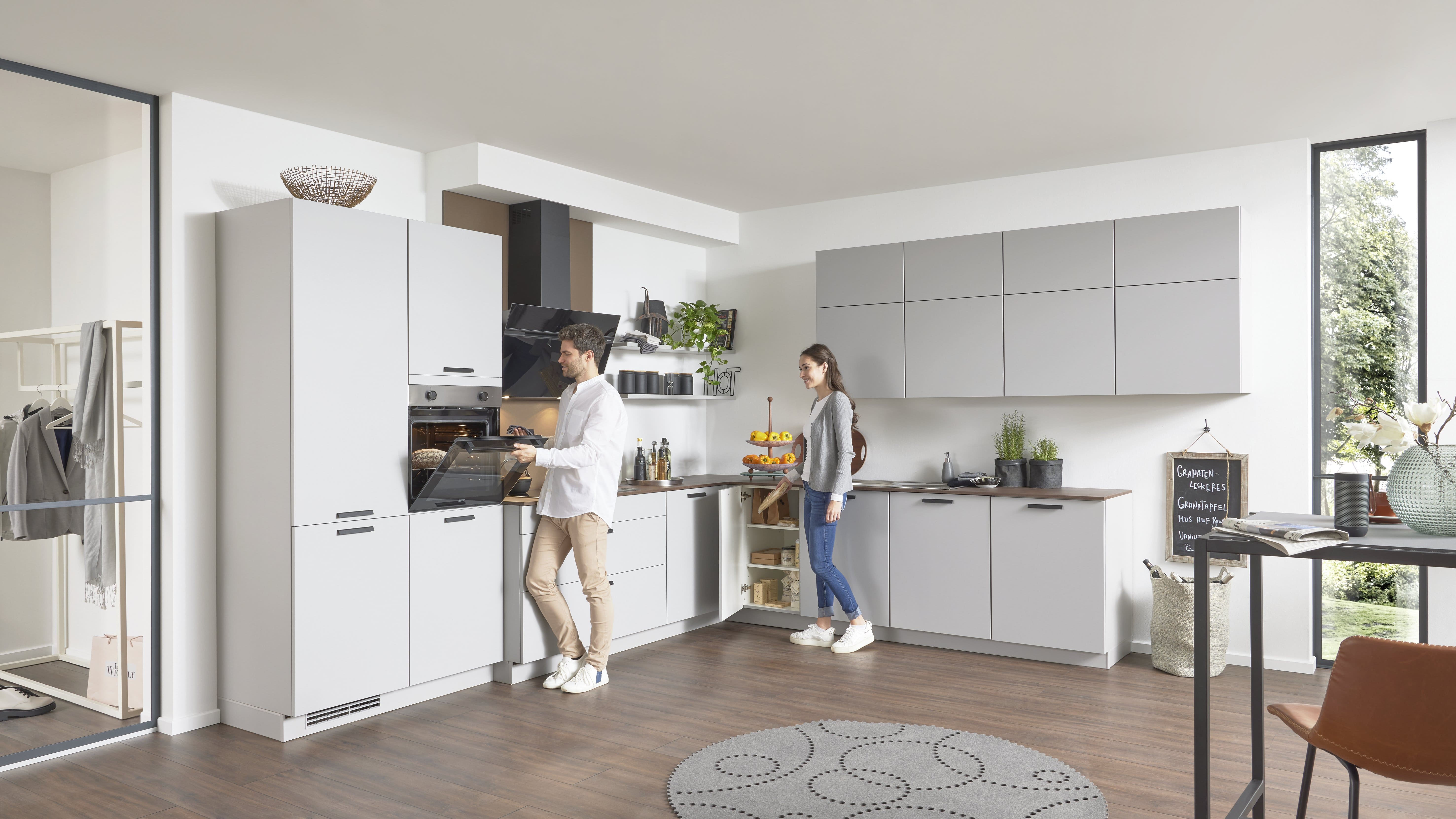 Weiße moderne Küche. Ein Mann schiebt etwas in den Ofen, eine Frau räumt was aus einem Schrank