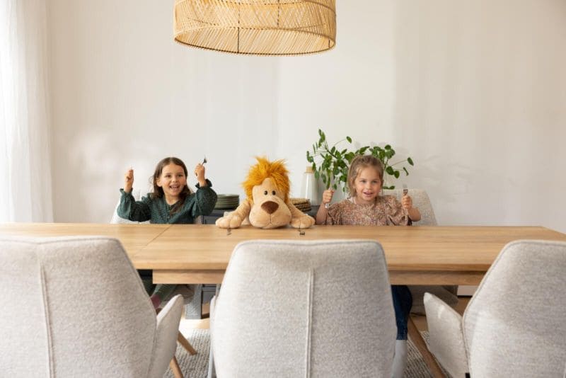 Zwei kleine Kinder, die am Tisch sitzen, zwischen ihnen hat ein Löwe Kuscheltier Platz genommen